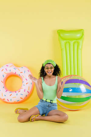 full length of cheerful african american woman sitting near inflatable ball, mattress and ring on...