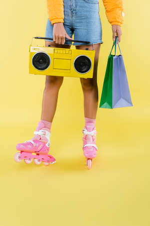 cropped view of african american woman in roller skates holding shopping bags and boombox on yellow