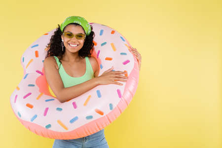positive african american young woman standing with inflatable ring isolated on yellow