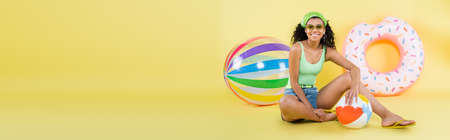 full length of happy african american woman sitting with beach ball on yellow, banner