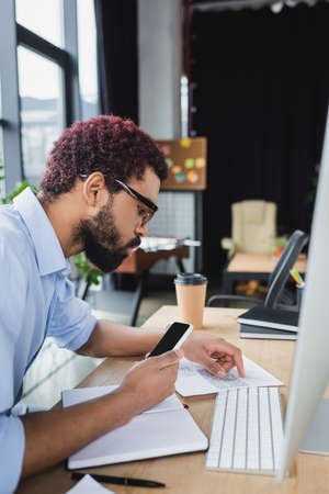 Side view of african american businessman holding smartphone near notebook and document in office