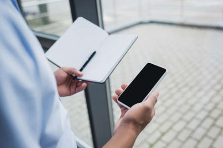 Cropped view of african american businessman holding cellphone with blank screen and notebook in...