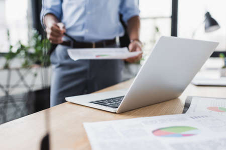 Cropped view of laptop near documents and blurred african american businessman in office