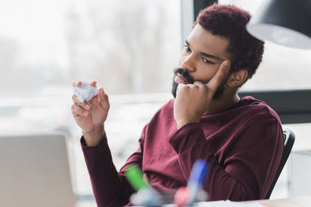 Focused african american businessman holding crumpled paper near laptop in office