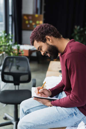 Side view of smiling african american businessman writing on notebook in officeの写真素材