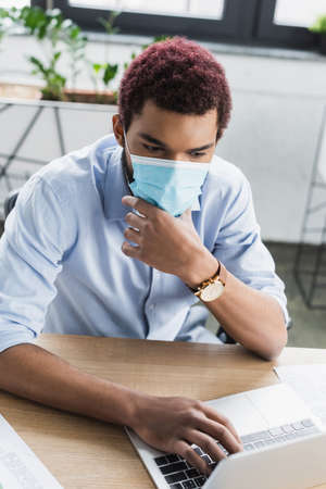 African american businessman in protective mask using laptop in officeの写真素材
