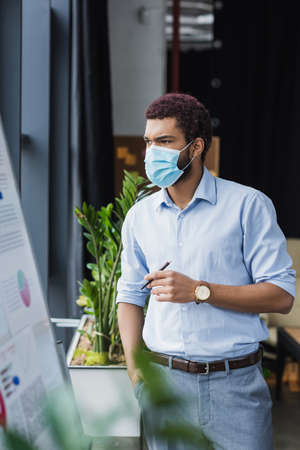African american businessman in medical mask looking at flip chart in officeの写真素材
