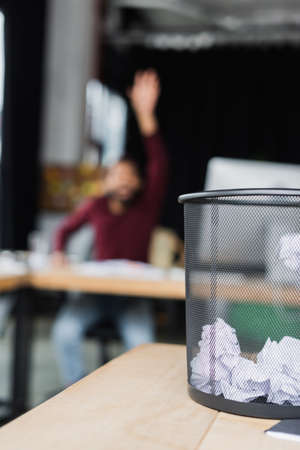 Trash can with crumpled paper on table near blurred african american businessman in officeの写真素材