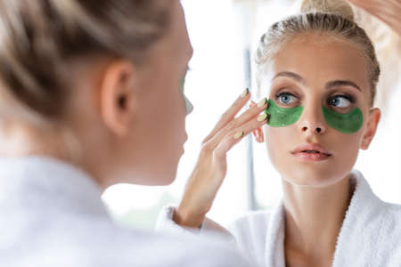 young woman in bathrobe applying green eye patches near mirrorの写真素材