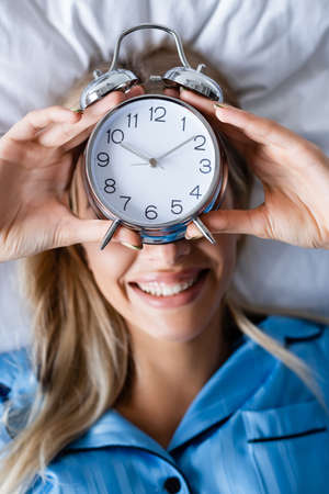 top view of cheerful woman holding retro alarm clock and covering face while lying on bedの写真素材