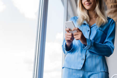 cropped view of happy young woman texting on cellphone near windowの写真素材