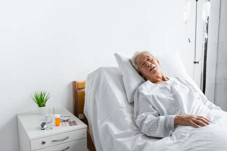 Ill asian patient lying on bed near pills and water in hospital wardの写真素材