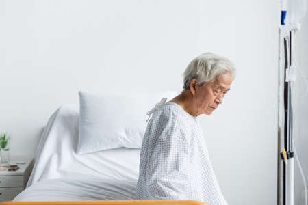Sad asian patient sitting on bed near intravenous therapy in hospital wardの写真素材