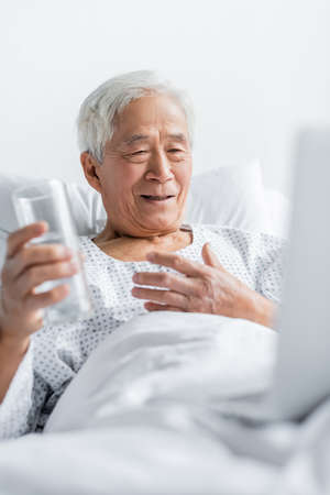 Smiling asian patient holding glass of water near blurred laptop on hospital bed