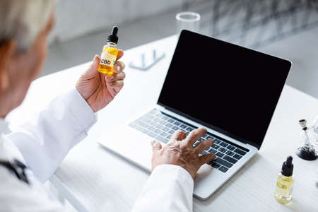 Blurred doctor holding cbd oil and using laptop with blank screen in clinicの写真素材
