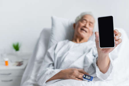 Smartphone in hand of blurred asian patient with pulse oximeter in hospital wardの写真素材