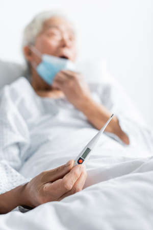 Electronic thermometer in hand on blurred elderly patient in hospital wardの写真素材