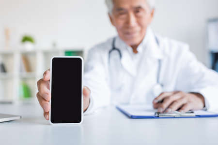 Blurred asian doctor holding smartphone with blank screen near clipboard in hospitalの写真素材