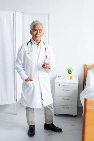 Senior asian doctor holding smartphone near bed in hospital wardの写真素材