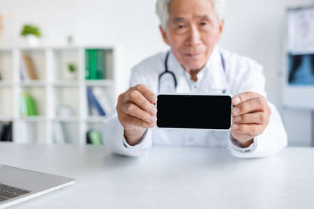 Smartphone with blank screen in hands of blurred asian doctor in hospital