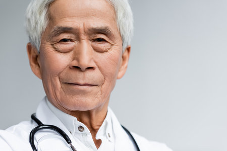 Portrait of senior asian doctor in white coat looking at camera isolated on greyの写真素材