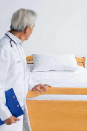 Blurred doctor with clipboard looking at bed in hospital wardの写真素材