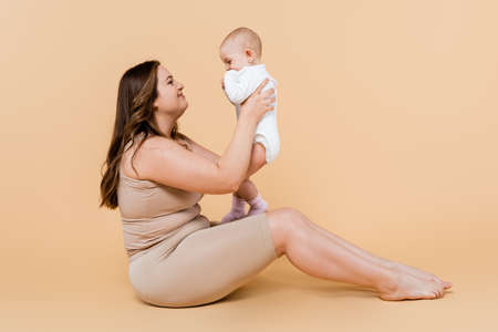 Side view of smiling plus size mother holding child while sitting on beige backgroundの写真素材