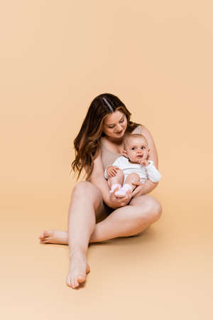 Smiling mother holding baby daughter while sitting on beige background