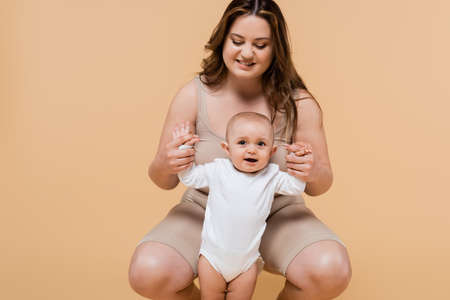 Positive plus size mother holding hands of cheerful daughter isolated on beige