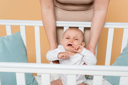 Plus size mother standing near crying baby daughter in crib isolated on beige