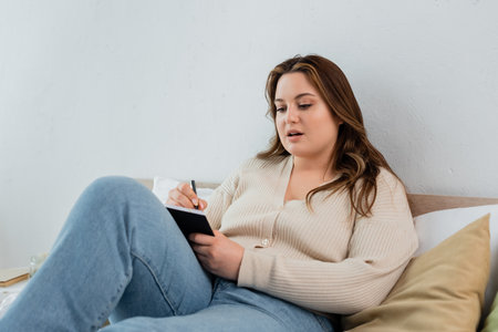Young plus size woman writing on notebook on bedの写真素材
