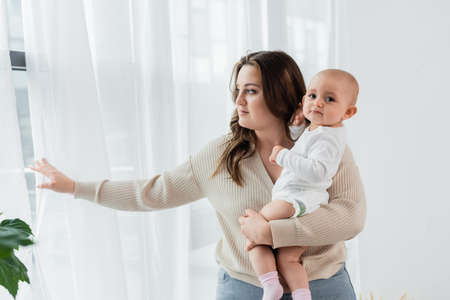 Pretty woman with overweight holding baby near curtains at homeの写真素材