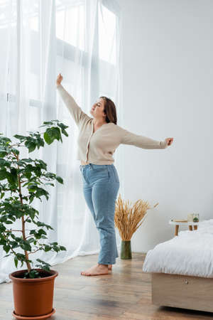 Young body positive woman stretching near window in bedroomの写真素材