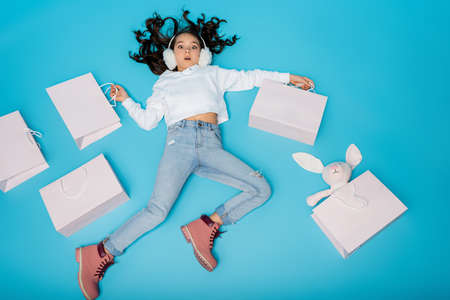 high angle view of shocked preteen girl in ear muffs lying near shopping bags and soft bunny on blue