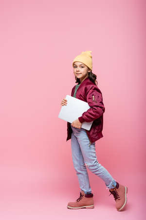 full length of stylish preteen girl in winter outfit posing with laptop on pinkの写真素材