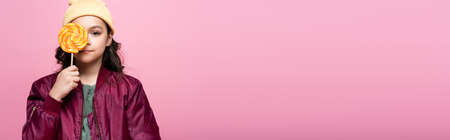 stylish preteen girl in winter outfit holding lollipop and covering eye isolated on pink, banner