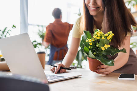 Smiling seller holding plant and using blurred laptop near smartphone in flower shopの写真素材