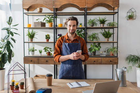 Florist holding smartphone near laptop and coffee in flower shopの写真素材