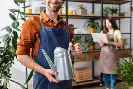 Smiling seller holding watering can near plants and colleague in flower shopの写真素材