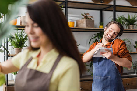 Cheerful florist talking on smartphone and writing on notebook near blurred colleague in shopの写真素材