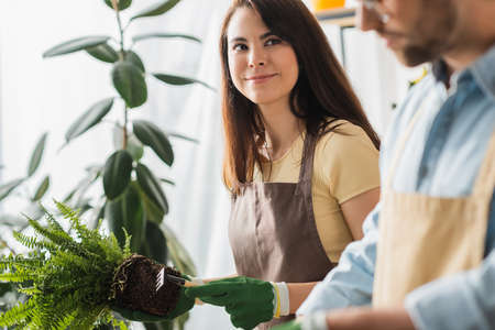 Florist in apron holding rake and plant near colleague in flower shopの写真素材