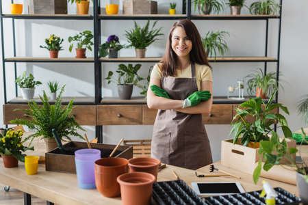 Smiling florist in gloves looking at camera near digital tablet and gardening tools in flower shopの写真素材