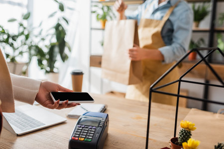 Cropped view of customer paying with smartphone near blurred florist with shopping bag in flower shopの写真素材