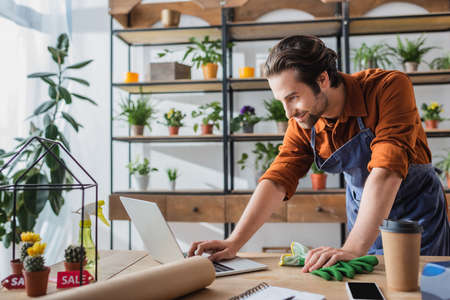 Young seller in apron using laptop near coffee to go and cacti in flower shopの写真素材