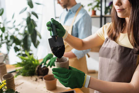 Cropped view of florist in apron pouring ground in flowerpot near blurred colleague in flower shopの写真素材