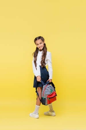 Smiling schoolgirl with backpack looking at camera on yellow backgroundの写真素材