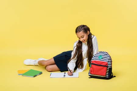 Smiling kid writing on notebook near backpack on yellow background