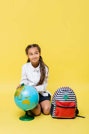 Smiling schoolkid sitting near globe and backpack on yellow backgroundの写真素材
