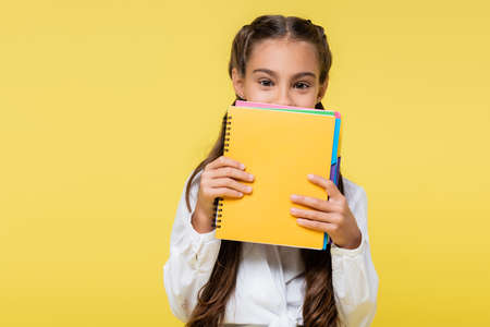 Schoolgirl covering face with notebooks isolated on yellowの写真素材