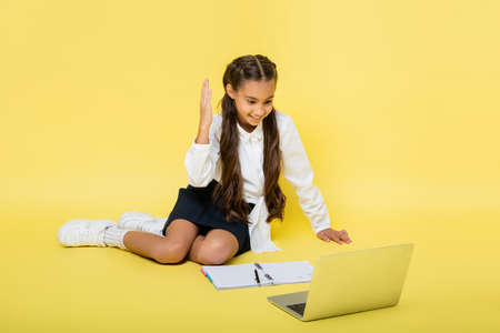 Smiling schoolkid raising hand during video call on laptop near notebook on yellow backgroundの写真素材